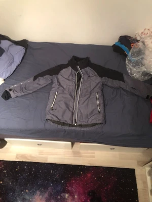 Grå vindjacka från Everest - Snygg grå vindjacka från Everest med svarta detaljer på axlar och ärmar. Jackan har två stora fickor med dragkedja framtill och en hög krage. Perfekt för blåsiga dagar och enkel att matcha med olika outfits.