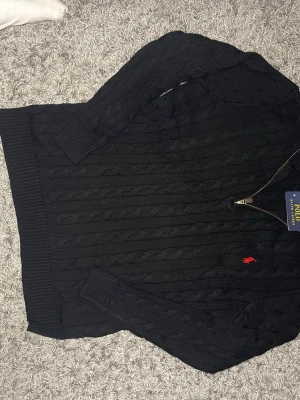 Svart kabelstickad tröja Polo Ralph Lauren - Kabelstickad svart tröja från Polo Ralph Lauren med halv dragkedja och röd broderad logga på bröstet. Tröjan har ribbade muddar och hög krage. Perfekt för dig som gillar klassisk och stilren design.