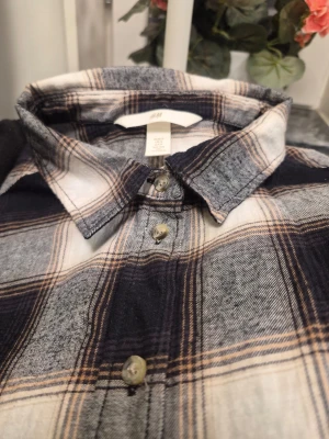 Rutig skjorta från H&M i bomull S - Säljer en klassisk rutig skjorta från H&M i svart, vit och beige. Skjortan har lång ärm, knappar framtill och mjukt bomullsmaterial. Perfekt för en avslappnad och stilren look.