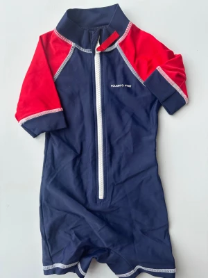 Marinblå och röd UV-dräkt Polarn O. Pyret - UV-dräkt från Polarn O. Pyret i marinblått med röda ärmar och vita kontrastsömmar. Dragkedja framtill och korta ben och ärmar. Perfekt för strand och pool. Tillverkad i stretchigt material som torkar snabbt.74/80
