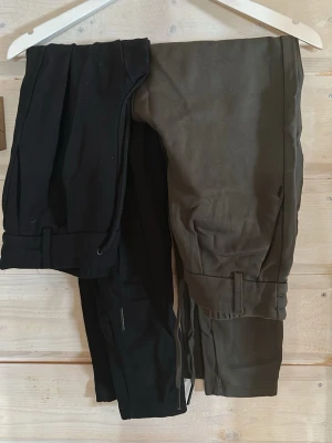 Svarta och gröna kostymbyxor med dragsko - Två par kostymbyxor i svart och mörkgrönt med raka ben och dragsko i midjan. Byxorna har en avslappnad passform och resår i midjan för extra komfort. Materialet ser ut att vara en mjuk bomullsblandning. Perfekta för en stilren och modern look. Båda par för 300kr