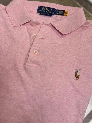 Ljusrosa piké från Polo Ralph Lauren - Säljer en ljusrosa pikétröja från Polo Ralph Lauren med klassisk krage, knappar framtill och den ikoniska broderade loggan på bröstet. Tröjan är i mjuk bomull och har korta ärmar. Perfekt för en clean och stilren look.