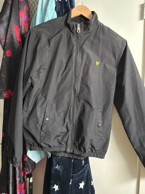Svart vindjacka från Lyle & Scott - Snygg svart vindjacka från Lyle & Scott med gul logga på bröstet. Jackan har dragkedja framtill, två fickor med tryckknappar och resår i midja och ärmslut. Tillverkad i lätt och tåligt material, perfekt för blåsiga dagar.