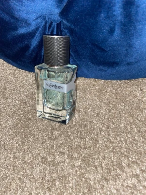 Yves Saint Laurent parfym herr - Stilren parfym från Yves Saint Laurent i en transparent, rektangulär glasflaska med ljusblå ton. Flaskan har en metallisk, mörkgrå kork med struktur. Märkets logga syns tydligt på framsidan. Perfekt för dig som gillar exklusiva dofter.