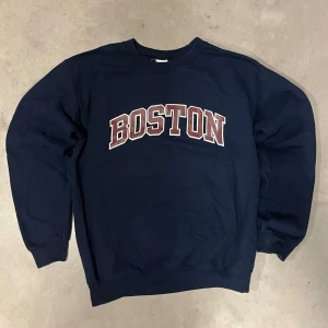 Mörkblå sweatshirt med Boston-tryck från Gildan - Snygg mörkblå sweatshirt med stort 'BOSTON'-tryck i vitt och vinrött på bröstet. Tröjan har rund halsringning, ribbade muddar vid ärmslut och nederkant samt en relaxed passform. Perfekt för dig som gillar college-stil. Materialet känns mjukt och bekvämt, troligen bomull eller bomullsblandning.