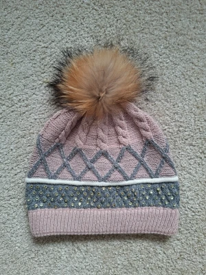 Rosa stickad mössa med pälsboll - Snygg rosa och grå stickad mössa med flätmönster och rutdetaljer. Mössan har en stor fluffig pälsboll i brunt och svart samt glittriga paljetter på den grå delen. Ribbad kant nedtill för extra komfort. Perfekt accessoar för kalla dagar.