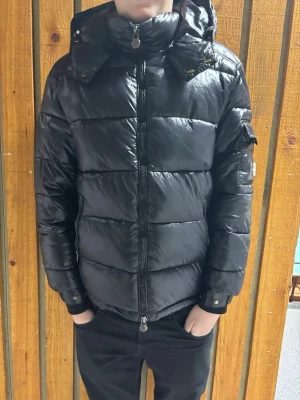 Svart dunjacka från Moncler - Snygg svart dunjacka från Moncler med glansig finish och klassisk puffer-design. Jackan har huva, hög krage, dragkedja framtill och Moncler-logga på ärmen. Perfekt för kalla vinterdagar och riktigt varm tack vare dunfyllningen.