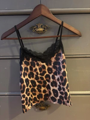 Leopardmönstrad linnetopp med spets - Säljer en leopardmönstrad linnetopp med tunna svarta axelband och svart spetskant upptill. Toppen har en croppad passform och är tillverkad i ett glansigt syntetmaterial som känns mjukt mot huden. Perfekt för dig som vill sticka ut med en edgy vibe.