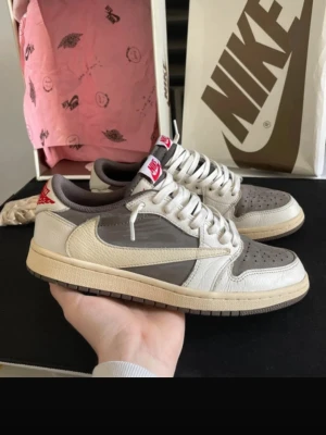Nike x Travis Scott Air Jordan 1 Low - Säljer ett par Nike x Travis Scott Air Jordan 1 Low med ikonisk omvänd Swoosh. Skorna är i beige, brunt och vitt läder med röda detaljer på hälen och tungan. Klassisk siluett med lågt skaft och vita skosnören. Perfekta för dig som gillar streetwear och unika collabs.