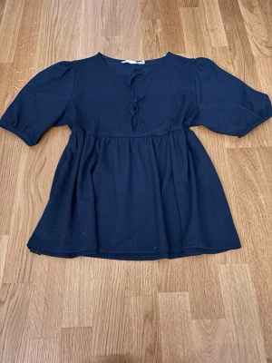 Marinblå blus med knyt framtill - Snygg marinblå blus med två dekorativa knyt framtill och lätt puffade ärmar. Blusen har en lös passform och är tillverkad i ett mjukt tyg som ger ett fint fall. Perfekt att styla med jeans eller kjol för en trendig look.