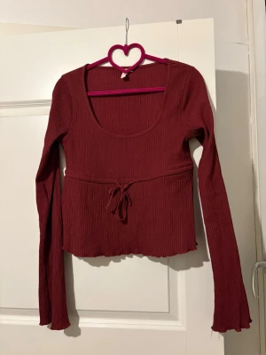 Vinröd ribbad topp med knytning - Trendig vinröd långärmad topp med ribbad struktur och söt knytning i midjan. Toppen har en croppad passform och en djup rundringning. Perfekt för dig som gillar en stilren men ändå lekfull look.