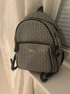 Mönstrad miniryggsäck från Guess - Snygg miniryggsäck från Guess i beige och svart med all-over logomönster. Väskan har en framficka med dragkedja, svarta detaljer och metallogo framtill. Tillverkad i konstläder och har justerbara axelband samt en dekorativ nyckelring.