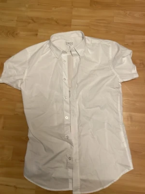 Vit kortärmad skjorta från Smog - Klassisk vit skjorta från Smog med korta ärmar och knappar framtill. Skjortan har en enkel och stilren design med krage och är tillverkad i ett lätt bomullsmaterial. Perfekt för en clean och fräsch look. Splitter ny