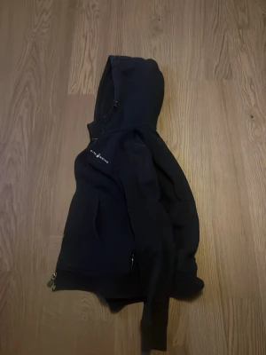 Svart hoodie från Sail Racing - Svart hoodie från Sail Racing med dragkedja och huva. Snygg logga på bröstet och praktiska fickor. Perfekt för en sportig och avslappnad stil. Materialet känns mjukt och bekvämt, passar till många tillfällen.