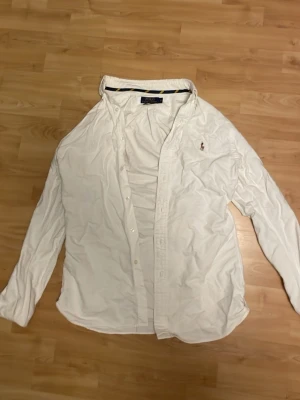 Vit skjorta från Polo Ralph Lauren - Klassisk vit skjorta från Polo Ralph Lauren med broderad logga på bröstet. Skjortan har lång ärm, knappar framtill och är tillverkad i mjuk bomull. Perfekt till en clean och stilren look. Den är använd 2 gånger så helt nyskick men lite skrynklig för den är tvättad. 