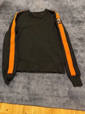 Parajumpers tröja  - Cool svart sweatshirt från Parajumpers med tydliga orange ränder längs båda ärmarna och en patch med logga på vänster ärm. Tröjan har rund halsringning och ribbade muddar vid ärmslut och nederkant. Perfekt för en sportig och avslappnad stil.