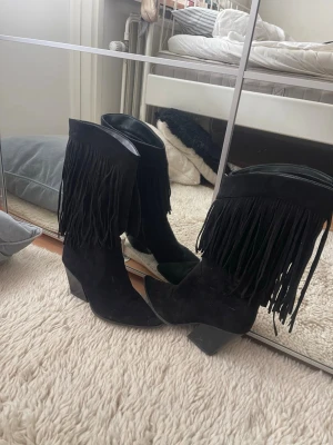 Svarta cowboy boots med fransar - Säljer ett par svarta cowboy boots i mocka-look med höga skaft och långa fransar längs kanten. Bootsens tå är spetsig och klacken är bred och stabil. Perfekta för dig som vill ha en western vibe i din outfit.
