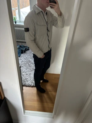 Vit vindjacka från Stone Island - Säljer en vit vindjacka från Stone Island med klassisk patch på ärmen. Jackan har dragkedja framtill, två bröstfickor med lock och en snygg krage. Perfekt för dig som gillar stilrena och sportiga plagg. Liten defekt på sista bilden.