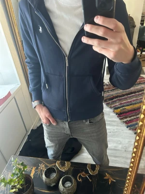 Mörkblå hoodie från Polo Ralph Lauren - Snygg mörkblå zip hoodie från Polo Ralph Lauren. Andvänd Max 3 gånger. Klassisk design med vit logga på bröstet. Tillverkad i mjukt bomullsmaterial som är super skönt. Perfekta plagget för dig som vill kombinera skönhet och stil. Tröjan är i storlek M men jag skulle säga att den passar de med storlek S
