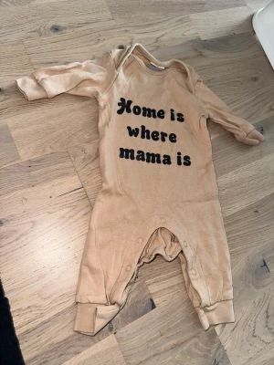 Beige babybody med text från KappAhl - Beige långärmad babybody från KappAhl med svart texten 'Home is where mama is' på framsidan. Praktiska tryckknappar i grenen för enkel påklädning. Mjuk och skön bomullskänsla, perfekt för de allra minsta.