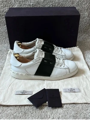 Valentino open i färgen kahki - Snygga vita sneakers från Valentino med ett brett olivgrönt band över mitten och coola nitar på hälen. Skorna har klassisk låg profil, vita skosnören och är tillverkade i läder. Perfekta för dig som vill ha en stilren men ändå unik look. De är en lite defekt på insidan av hälen och så är de målade längst fram för en längre hållbarhet.