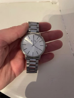 Silverfärgad klocka från Armani Exchange - Stilren klocka från Armani Exchange med silverfärgad länkarmband och rund urtavla. Urtavlan är vit med minimalistiska svarta markeringar och AX-logga. Klockan har en klassisk och clean look som passar till det mesta.