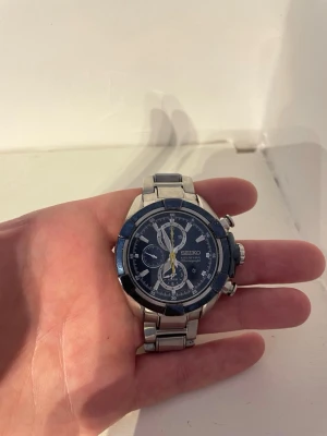 Seiko blå kronograf herrklocka - Snygg Seiko herrklocka med blå urtavla och silverfärgat armband i metall. Klockan har kronograf-funktion, tydliga index och flera små urtavlor. Robust design med sportig känsla och detaljer i gult på visarna. Perfekt accessoar för dig som gillar stilrena klockor.