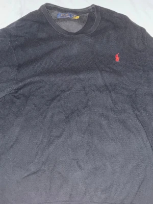 Svart stickad tröja Polo Ralph Lauren - Snygg svart stickad tröja från Polo Ralph Lauren med rund halsringning och den klassiska röda logotypen broderad på bröstet. Tröjan har en enkel och stilren design med ribbade muddar vid ärmslut och nederkant. Perfekt för en avslappnad men ändå trendig look.