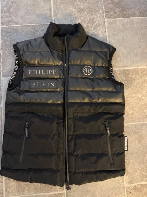 Svart dunväst från Philipp Plein - Snygg svart dunväst från Philipp Plein med hög krage och dragkedja framtill. Västen har två fickor med dragkedja och tydliga logotexter på bröstet samt logga på vänster sida. Materialet är glansigt och quiltat, vilket ger en exklusiv känsla.    Texten bakom dragkedjan börjar gå bort men inget som syns när den är stängd. 