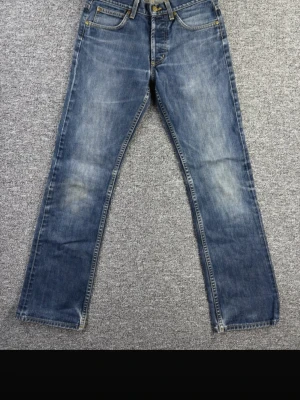 Blå raka jeans med slitningar - Ett par klassiska blå jeans med raka ben och snygga slitningar på framsidan. Jeansen har fem fickor, guldfärgade sömmar och knappgylf. Materialet är denim i bomull och passformen är avslappnad men ändå stilren.