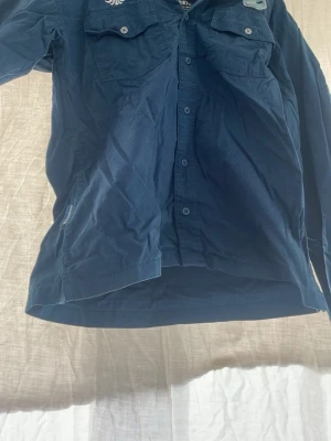 Mörkblå scoutskjorta med bröstfickor(knivmärke som man kan ta bort) - Klassisk mörkblå scoutskjorta från Scouterna med två bröstfickor och knappar framtill. Skjortan har lång ärm och är tillverkad i bomull. Perfekt för scoutaktiviteter eller som en cool vardagslook.