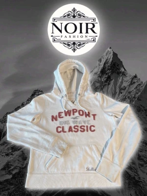 Vit hoodie från Hollister med tryck - Vit hoodie från Hollister med röd text 'NEWPORT BIG WAVE CLASSIC' på bröstet och broderad logga vid fickan. Klassisk känguruficka och dragsko i huvan. Perfekt för en avslappnad och cool stil.