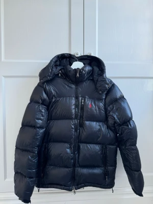 Svart dunjacka från Polo Ralph Lauren - Svart dunjacka från Polo Ralph Lauren med blank finish, avtagbar huva och flera dragkedjeförsedda fickor. Klassisk puffer-modell med röd logotyp på bröstet. Perfekt för kalla vinterdagar och riktigt snygg allmänt.