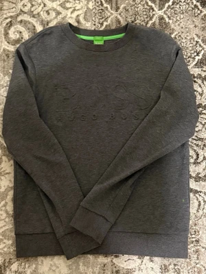 Mörkgrå sweatshirt från Hugo Boss - Mörkgrå sweatshirt från Hugo Boss med diskret broderad logga på bröstet. Tröjan har rund halsringning, ribbade muddar vid ärmslut och nederkant samt en stilren, enkel design. Perfekt för en avslappnad och snygg look.