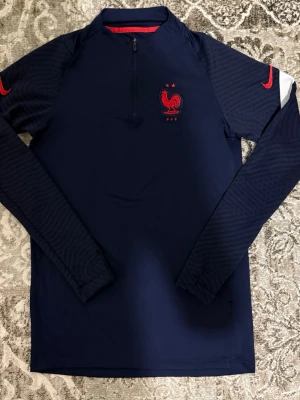Frankrike tröja - Mörkblå långärmad träningströja från Nike med FFF-logga och röd tupp på bröstet. Tröjan har halv dragkedja framtill, röda Nike-loggor på ärmarna och är tillverkad i ett lätt funktionsmaterial.