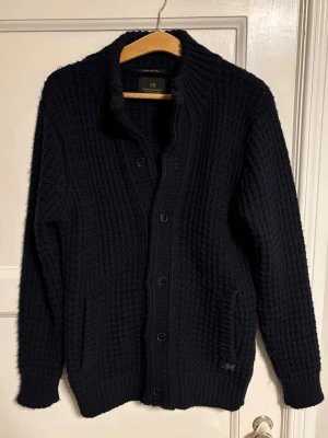 Mörkblå stickad kofta från Scotch & Soda - Mörkblå stickad kofta från Scotch & Soda med grov struktur, knappar framtill och två fickor.         Storlek L.                                                                              Hör av er för mer information/bilder
