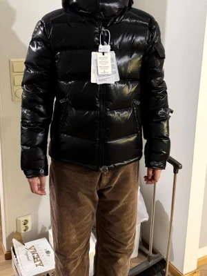 Svart dunjacka från Moncler Size 1 - Säljer en glansig svart dunjacka från Moncler med huva och dragkedja framtill. Jackan har två snedställda fickor med dragkedja och en ficka på ärmen med Moncler-logga. Puffig modell som håller dig varm under kalla dagar. Size 1- XS-S. Sell with garment bag. Size 1-3 are all available. 