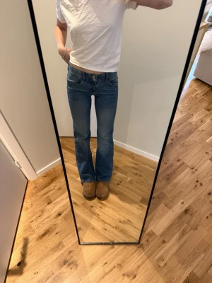 Blå bootcut jeans med låg midja - Säljer ett par klassiska blå bootcut jeans med låg midja och fem fickor. Jeansen har en snygg tvätt och är tillverkade i ett stretchigt denimtyg som sitter skönt. Perfekta för dig som gillar en avslappnad men trendig look.