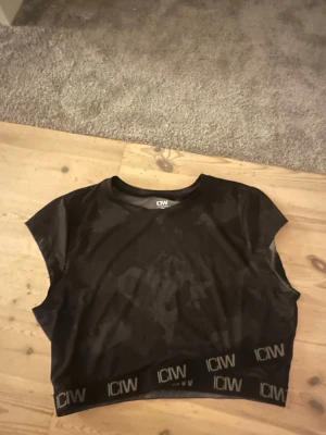 Svart croppad topp från ICANIWILL - Svart croppad topp från ICANIWILL med diskret camouflage-mönster. Toppen har korta ärmar och bred resår med logga nertill. Tillverkad i ett stretchigt syntetmaterial som passar perfekt till träning.