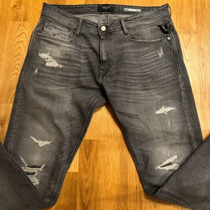 Grå slitna jeans från Replay Rocco - Säljer ett par grå jeans från Replay, modell Rocco Comfort Fit. Jeansen har slitna detaljer och en klassisk femficksdesign. De har raka ben och en snygg tvättad look med ljusare partier och slitningar på låren och knäna. Priset är inte hugget i sten och det är bara att höra av dig vid minsta fundering:).