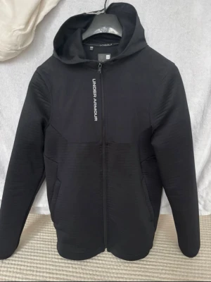 Svart vindjacka från Under Armour - Säljer en svart vindjacka från Under Armour med dragkedja framtill och reflekterande logga på bröstet. Jackan har två sidofickor och en skön huva. Materialet är mjukt och stretchigt, perfekt för träning eller chill. Enkel och stilren design.