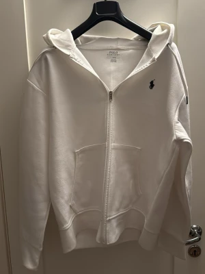 Vit zip hoodie från Polo Ralph Lauren - Klassisk vit hoodie med dragkedja från Polo Ralph Lauren. Hoodien har två stora fickor framtill, justerbar huva och den ikoniska lilla mörkblå loggan broderad på bröstet. Tillverkad i mjuk bomull för en skön och avslappnad look.