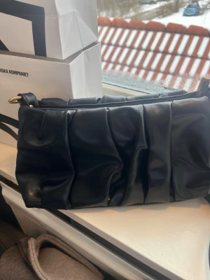 Svart quiltad handväska med axelrem - Trendig svart handväska i quiltad design med mjukt, veckat konstläder och guldfärgade metalldetaljer. Väskan har en avtagbar axelrem och dragkedja upptill. Perfekt för dig som gillar en stilren men ändå unik look.