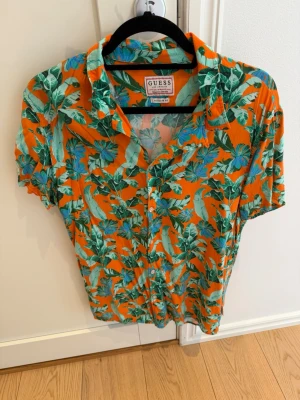 GUESS x J Balvin Tropical Print Resort Shirt – Strl S – Kanonskick - Färgstark och iögonfallande kortärmad skjorta från GUESS med ett tropiskt mönster i härliga orange, gröna och blå toner. En perfekt skjorta för semestern, sommarfester, stranden eller för dig som vill addera färg och personlighet till din stil.  ✔ Märke: GUESS ✔ Storlek: S ✔ Passform: Regular Fit ✔ Kortärmad Resort/Hawaiiskjorta ✔ Tropiskt palmbladsmönster i livfulla färger ✔ Design inspirerad av GUESS semesterkollektion och Guess x J Balvin-serien ✔ Kanonskick  Kommer från ett djur- och rökfrit