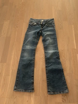 Blå bootcut jeans med dubbla knappar - Säljer ett par blå bootcut jeans från Gina Tricot med dubbla silverknappar i midjan och coola slitningar. Jeansen har klassiska fickor fram och bak samt en liten ficka med lock. Perfekta för dig som gillar retrovibbar och vill ha en snygg siluett.