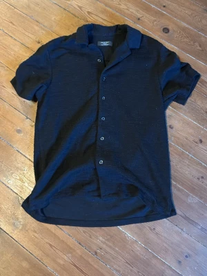 Svart kortärmad skjorta - Stilren svart skjorta med korta ärmar och klassisk krage. Skjortan har knappar framtill och är tillverkad i ett mjukt, strukturerat material som känns skönt mot huden. Perfekt för en avslappnad och trendig look.