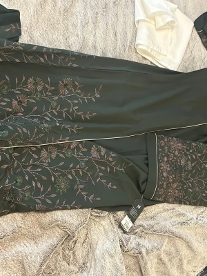 Blommig svart  med brodyr - Elegant svart abaya  med detaljerad blombroderi i grönt och lila. Lång modell med bälte i midjan och kontrasterande vita passpoaler längs kanterna. Perfekt för en lyxig och avslappnad känsla hemma.