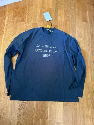 Blå långärmad topp Acne Studios - Mörkblå långärmad topp från Acne Studios med tryck 'Acne Studios STOCKHOLM 1996' på bröstet. Klassisk rund halsringning och relaxed fit, perfekt för en stilren och enkel look. Priset kan självklart diskuteras!