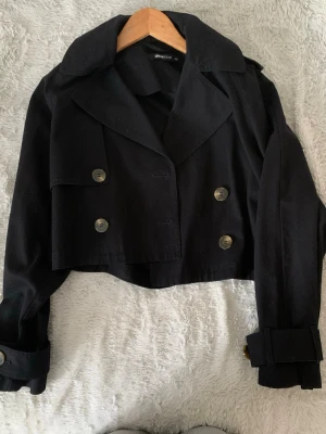 Kort svart trenchcoat från gina tricot - Snygg svart oversized trenchcoat i kort modell från gina tricot i storlek xxs Jackan har dubbelknäppning med stora knappar, bred krage och justerbara ärmslut med spänne. Perfekt för dig som gillar en trendig och stilren look. Har använt 2 gånger innan men är i mycket bra skicks 