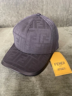 Mörkgrå Fendi keps med FF-mönster - Snygg mörkgrå keps från Fendi med det klassiska FF-logomönstret över hela kepsen. Skärmen har en stilren kant i läderimitation och baktill finns ett justerbart spänne i svart läder med Fendi Roma-inpräglad logga. Perfekt för dig som gillar exklusiva accessoarer.